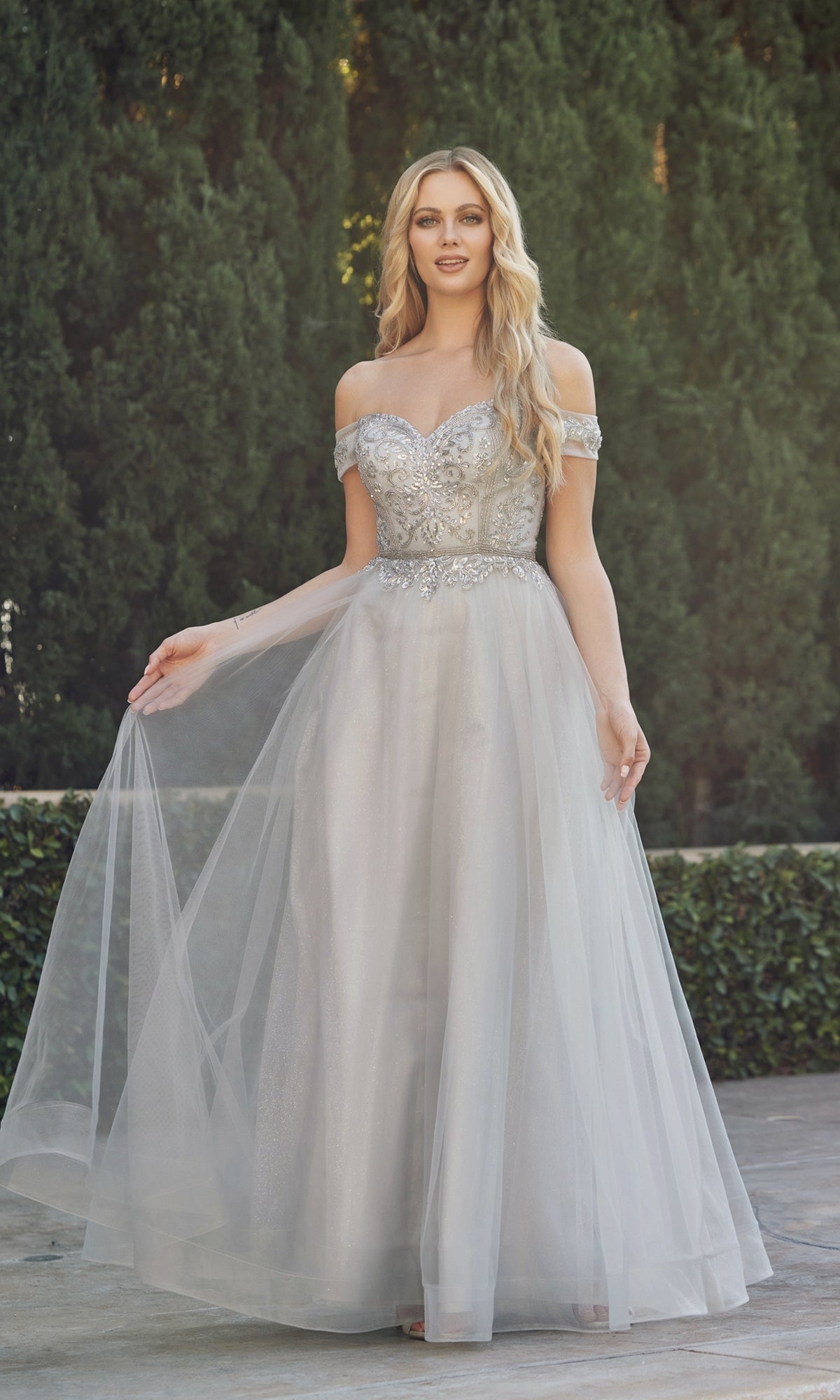 A woman stands outdoors before lush green hedges, wearing the Juliet 287 Long Sweetheart Prom Ball Gown—a silver off-the-shoulder dress with a beaded corset bodice and flowing tulle skirt.